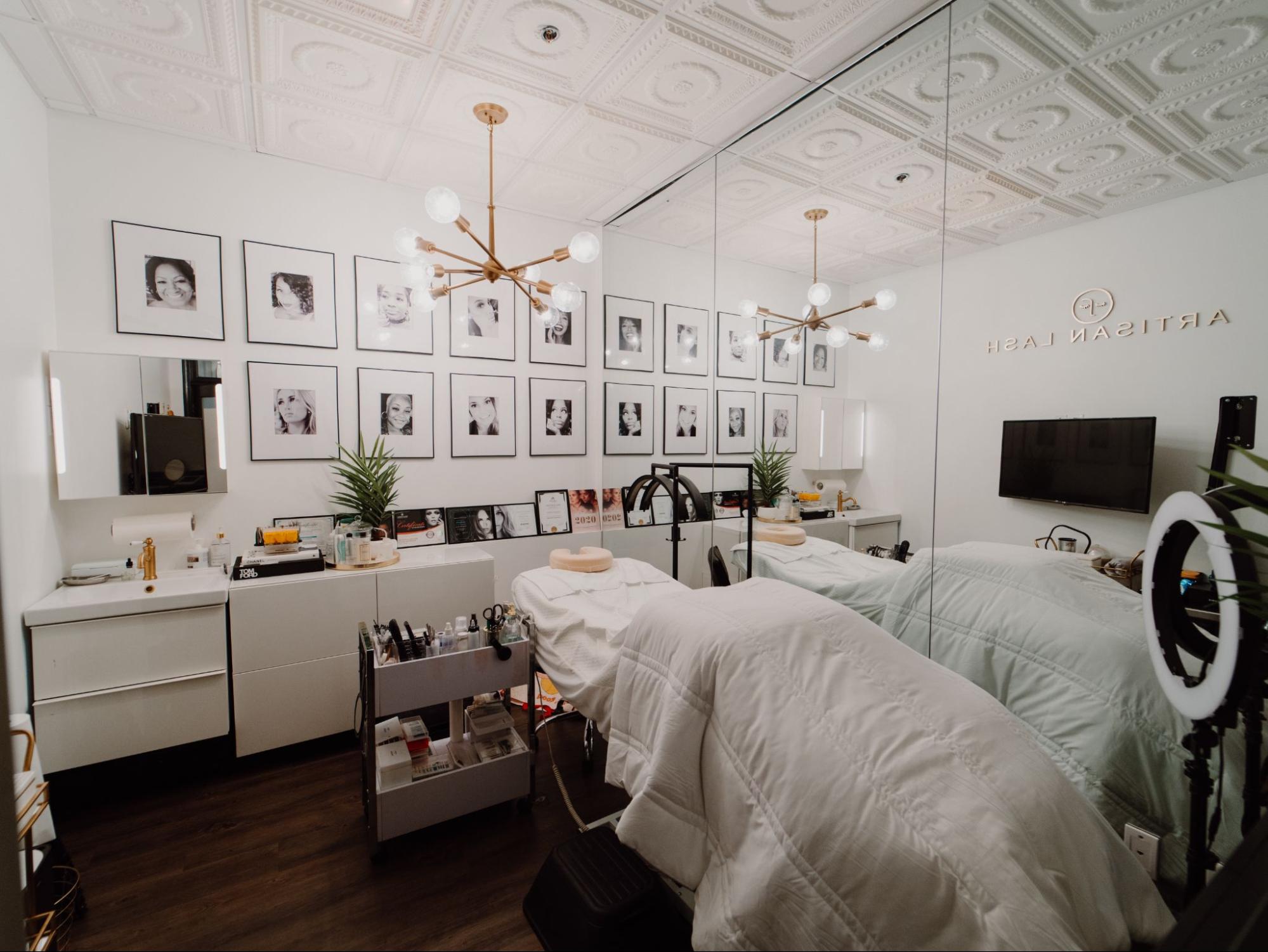 Salon Studio Rental - Modern and Elegant Private Beauty Suite Modern salon studio rental with treatment bed, ring light, large mirror, stylish chandelier, and framed photos on wall.